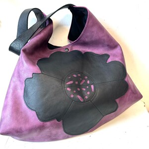 Puede incluir: Un bolso tote de cuero morado con un dise&ntilde;o de flor de cuero negro. La flor tiene un patr&oacute;n recortado.