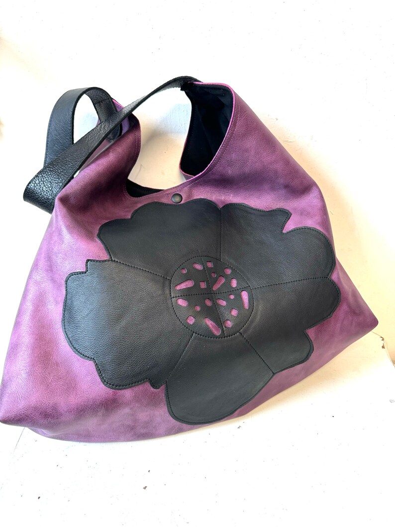 Puede incluir: Un bolso tote de cuero morado con un dise&ntilde;o de flor de cuero negro. La flor tiene un patr&oacute;n recortado.