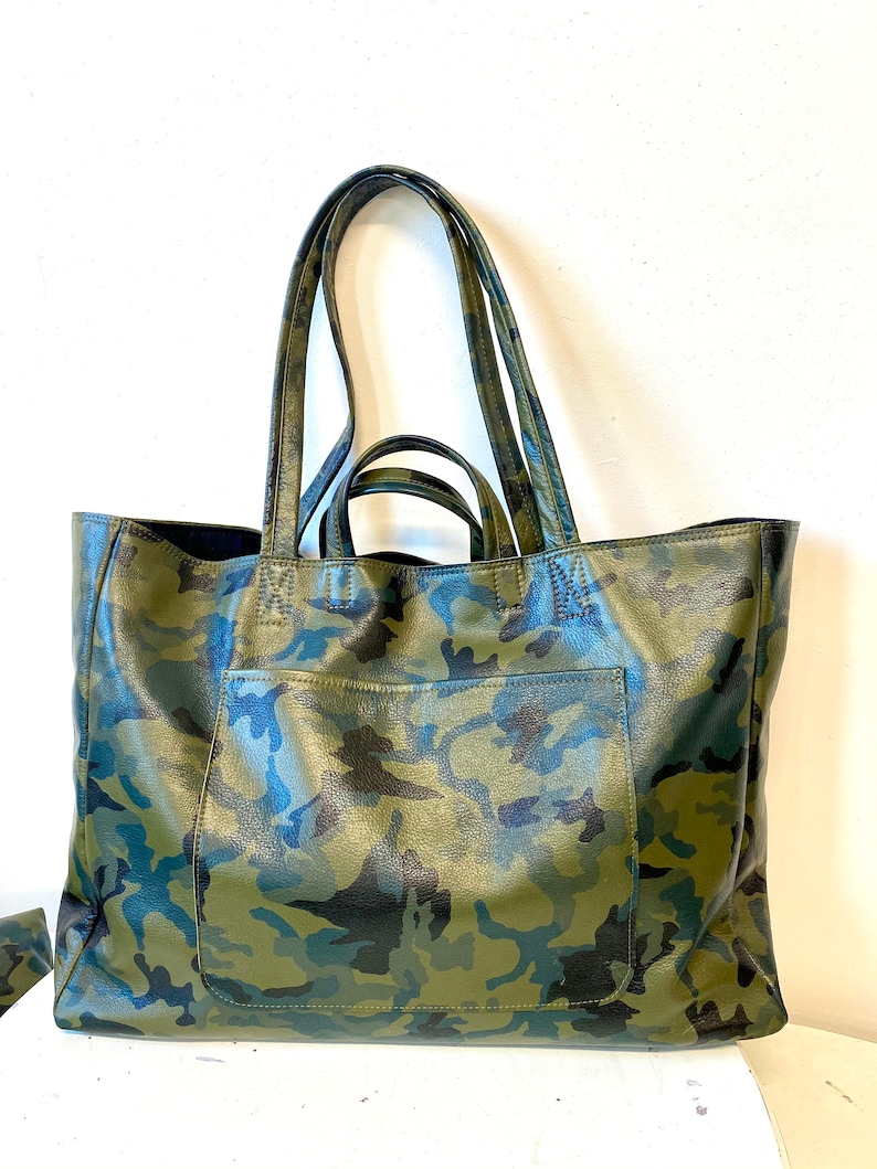 May include: A large tote bag with a camouflage pattern in shades of green, blue, and black. The bag has two long handles and a front pocket. The bag is made of a textured material.