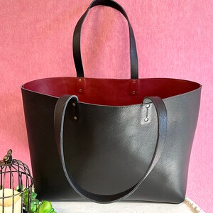 May include: A large black tote bag with a burgundy interior. The bag has two long black handles and is made of textured leather-like material. The bag is sitting on a white surface against a pink background.