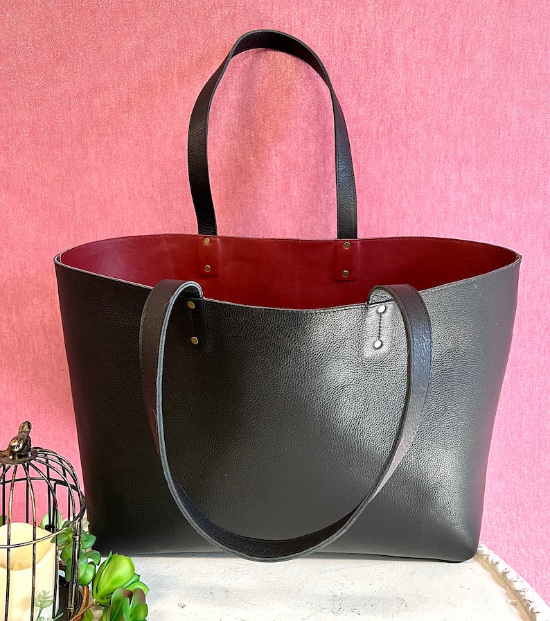 May include: A large black tote bag with a burgundy interior. The bag has two long black handles and is made of textured leather-like material. The bag is sitting on a white surface against a pink background.