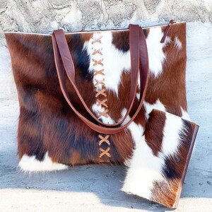 K&ouml;nnte beinhalten: Eine braune und wei&szlig;e Kuhfell-Tragetasche mit braunen Lederriemen und einer passenden kleineren Tasche. Die Tasche hat einen Rei&szlig;verschluss und ein dekoratives Steppdesign.