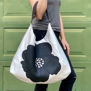 Puede incluir: Un bolso tote de cuero plateado con un gran dise&ntilde;o de flor negra. El bolso tiene una sola correa y lo lleva una persona.