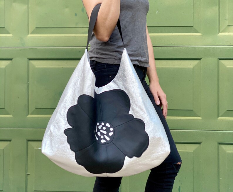 Puede incluir: Un bolso tote de cuero plateado con un gran dise&ntilde;o de flor negra. El bolso tiene una sola correa y lo lleva una persona.