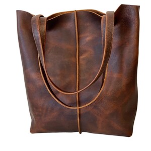May include: A brown leather tote bag with a distressed finish and a single stitch detail down the center. The bag has two long straps.