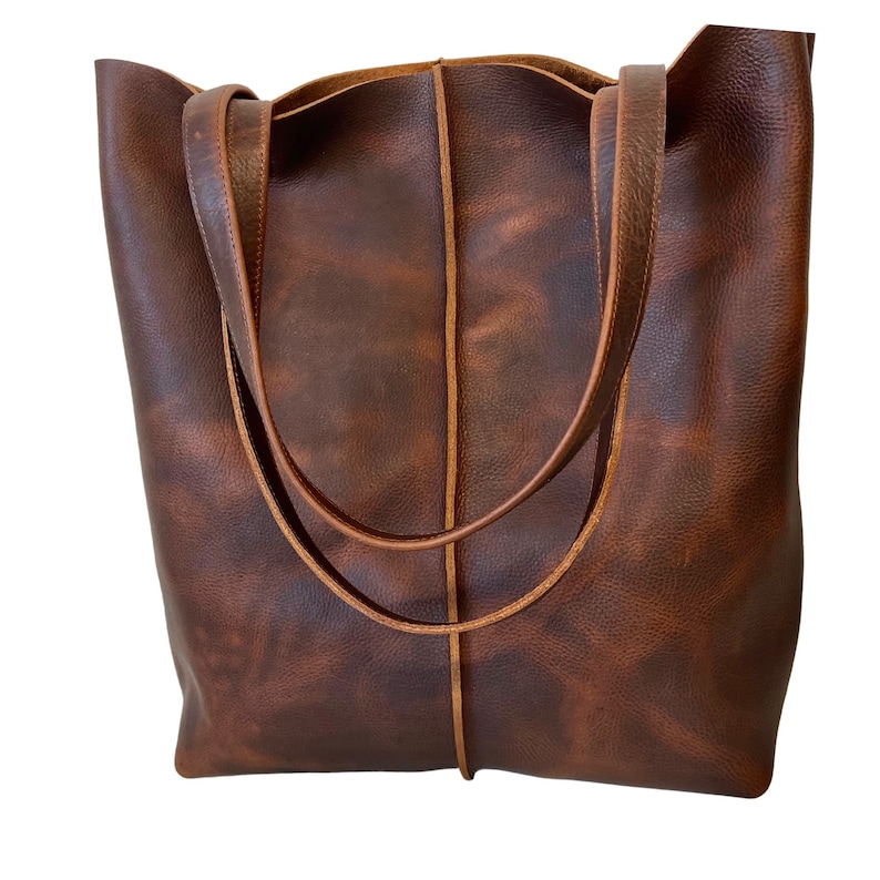 May include: A brown leather tote bag with a distressed finish and a single stitch detail down the center. The bag has two long straps.
