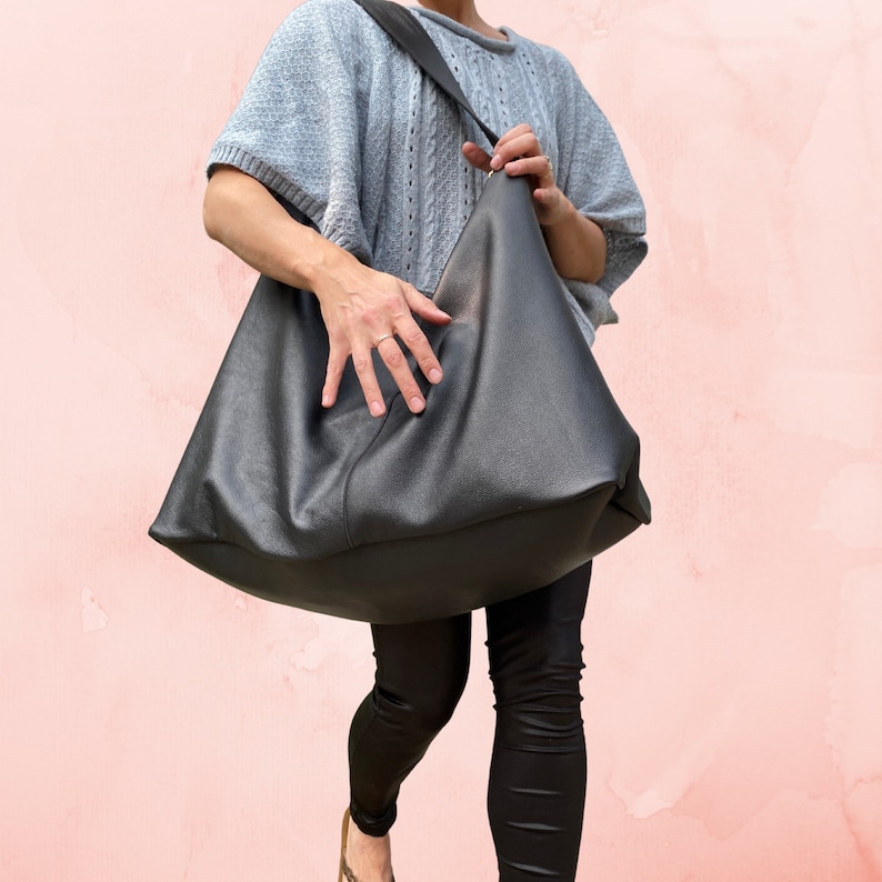 May include: A large black leather tote bag with a long strap. The bag is being held by a person wearing black leggings and a light gray sweater.