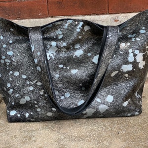 May include: A large black and white tote bag with a cowhide pattern. The bag has two black straps and a silver metallic finish.