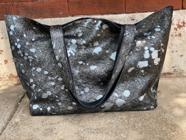 May include: A large black and white tote bag with a cowhide pattern. The bag has two black straps and a silver metallic finish.