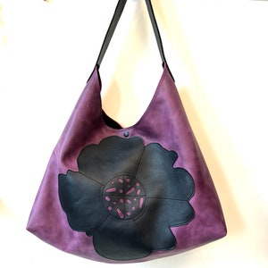 Puede incluir: Un bolso tote de cuero morado con un dise&ntilde;o de flor negra. La flor tiene un dise&ntilde;o de corte en el centro.