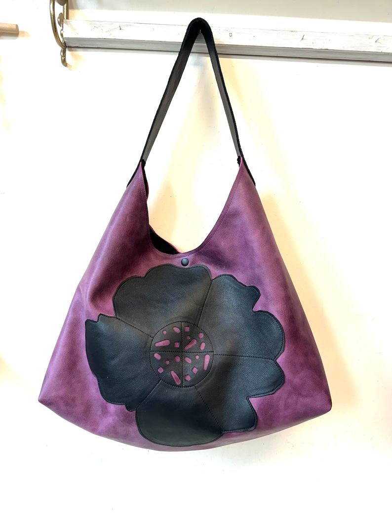 Puede incluir: Un bolso tote de cuero morado con un dise&ntilde;o de flor negra. La flor tiene un dise&ntilde;o de corte en el centro.