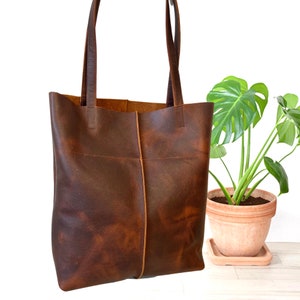 May include: A brown leather tote bag with a wide, rectangular shape and two long handles. The bag has a distressed look and is made of a thick, durable leather.