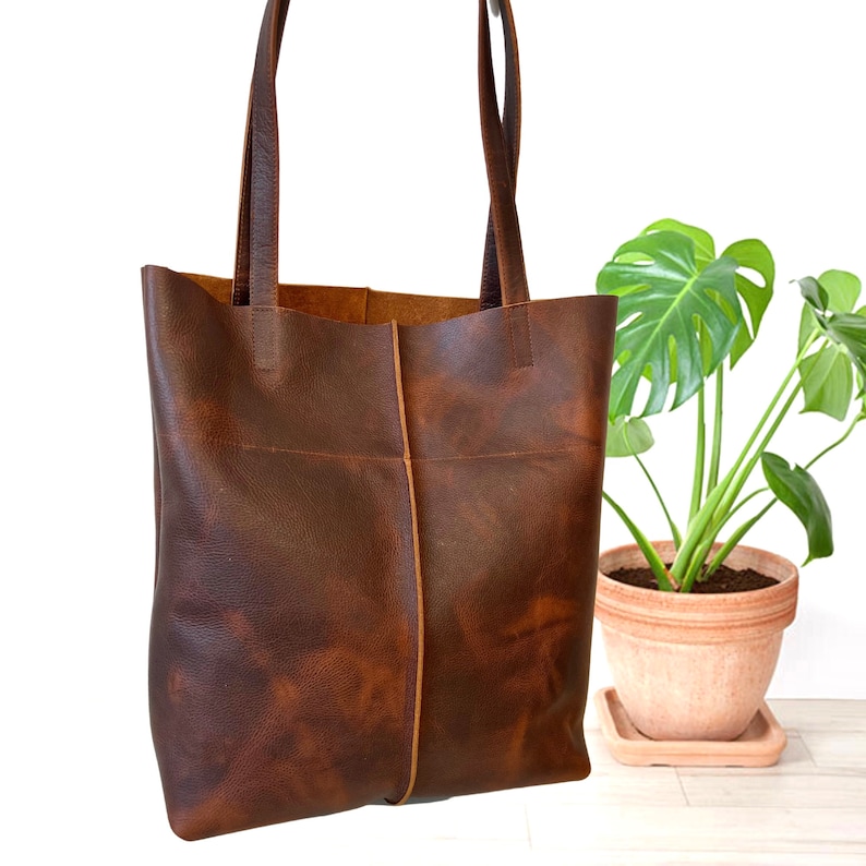 May include: A brown leather tote bag with a wide, rectangular shape and two long handles. The bag has a distressed look and is made of a thick, durable leather.