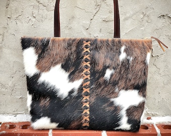 May include: A brown, black, and white cowhide tote bag with brown leather straps and a braided leather detail down the center. The bag has a zipper closure.