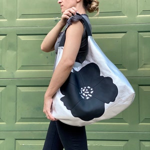 Puede incluir: Un bolso tote de cuero plateado con un gran dise&ntilde;o de flor negra. La flor tiene un centro blanco con puntos negros.