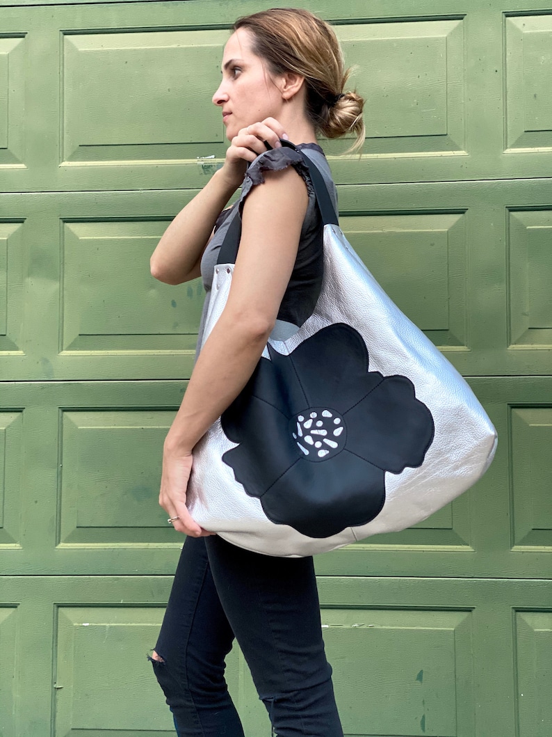 Puede incluir: Un bolso tote de cuero plateado con un gran dise&ntilde;o de flor negra. La flor tiene un centro blanco con puntos negros.