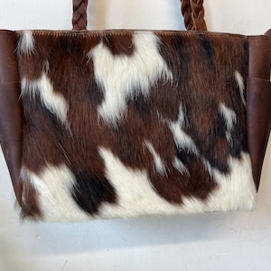 Op de afbeelding: Een tote bag van koeienhuid met een bruin, wit en zwart vlekkenpatroon. De tas heeft bruine leren zijkanten en gevlochten leren handvatten. Twee zakken zijn zichtbaar aan de voorkant van de tas.