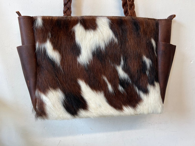 Op de afbeelding: Een tote bag van koeienhuid met een bruin, wit en zwart vlekkenpatroon. De tas heeft bruine leren zijkanten en gevlochten leren handvatten. Twee zakken zijn zichtbaar aan de voorkant van de tas.