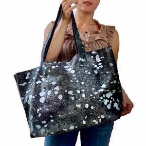 May include: A large black and white tote bag with a cowhide pattern. The bag has two black straps and is being held by a person.