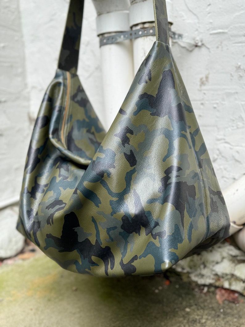 May include: A green, black, and blue camouflage-patterned leather shoulder bag. The bag has a long strap and a zipper closure.