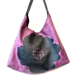 Puede incluir: Un bolso tote de cuero morado con un dise&ntilde;o de flor de cuero negro. La flor tiene un dise&ntilde;o de corte en el centro.