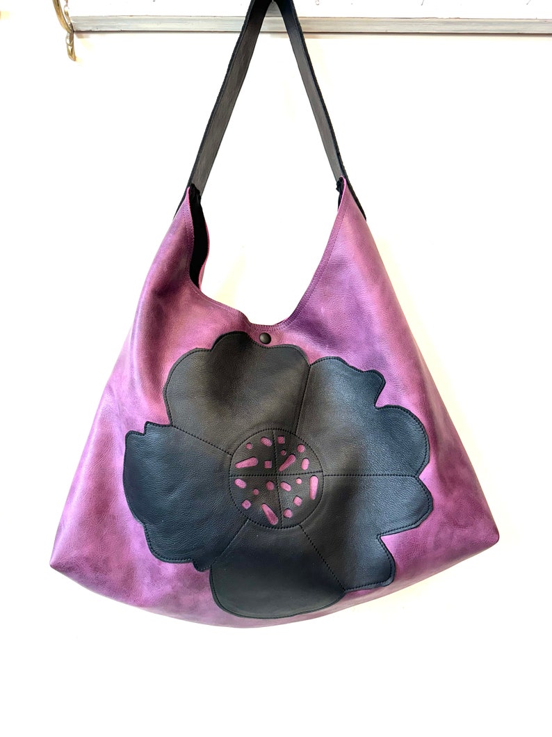 Puede incluir: Un bolso tote de cuero morado con un dise&ntilde;o de flor de cuero negro. La flor tiene un dise&ntilde;o de corte en el centro.