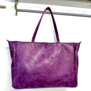 May include: A large purple leather tote bag with two handles. The bag is hanging on a white shelf.