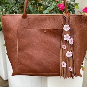 Op de afbeelding: Een bruine leren tote bag met een voorvak en handvatten aan de bovenkant. Een decoratieve kwast met roze en witte bloemaccenten hangt aan het handvat. De tas heeft een ritssluiting en een goudkleurige rits.