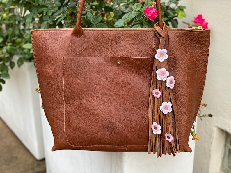 Op de afbeelding: Een bruine leren tote bag met een voorvak en handvatten aan de bovenkant. Een decoratieve kwast met roze en witte bloemaccenten hangt aan het handvat. De tas heeft een ritssluiting en een goudkleurige rits.