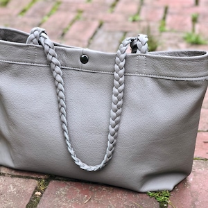 Op de afbeelding: Een grijze leren tote bag met gevlochten handvatten. De tas heeft een rechthoekige vorm met een knoopsluiting en een gestikte bovenrand. De dikke, gevlochten handvatten geven de tas een stijlvol detail.