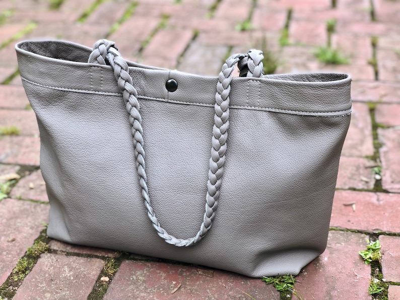 Op de afbeelding: Een grijze leren tote bag met gevlochten handvatten. De tas heeft een rechthoekige vorm met een knoopsluiting en een gestikte bovenrand. De dikke, gevlochten handvatten geven de tas een stijlvol detail.