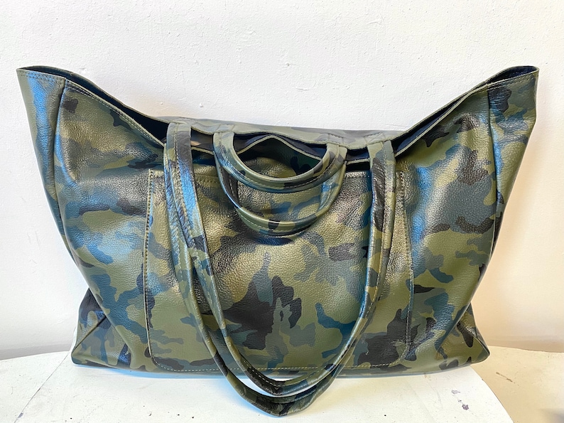May include: Large tote bag with a camouflage pattern in shades of green, black, and brown. The bag has two handles and a spacious interior. The bag is made of a textured material.