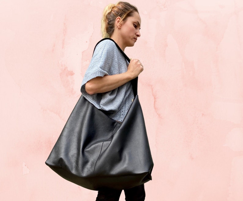 May include: A large black leather tote bag with a gold zipper. The bag is being carried by a person wearing a gray sweater.