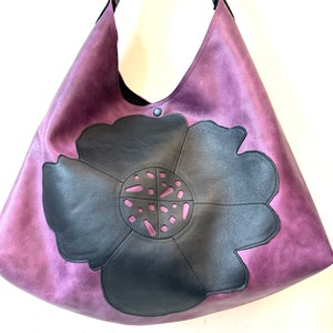 Puede incluir: Un bolso tote grande de cuero morado con un dise&ntilde;o de flor de cuero negro. La flor tiene un patr&oacute;n de corte en el centro.