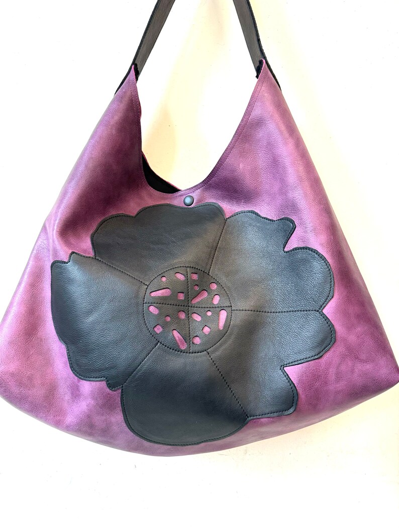 Puede incluir: Un bolso tote grande de cuero morado con un dise&ntilde;o de flor de cuero negro. La flor tiene un patr&oacute;n de corte en el centro.