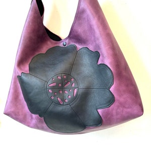 Puede incluir: Un bolso tote grande de cuero morado con un dise&ntilde;o de flor de cuero negro. La flor tiene un patr&oacute;n recortado.