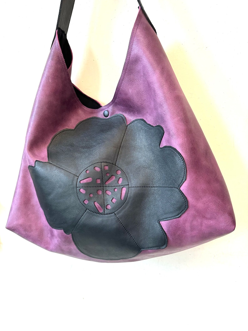 Puede incluir: Un bolso tote grande de cuero morado con un dise&ntilde;o de flor de cuero negro. La flor tiene un patr&oacute;n recortado.