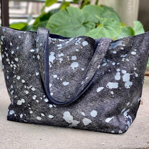 May include: A black and white cowhide tote bag with a black leather trim. The bag has a large, open top and two handles.