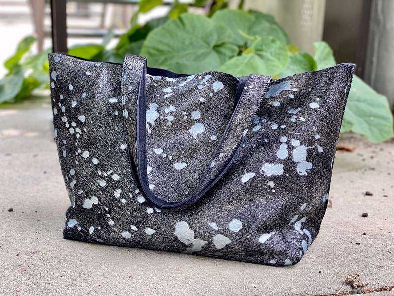 May include: A black and white cowhide tote bag with a black leather trim. The bag has a large, open top and two handles.
