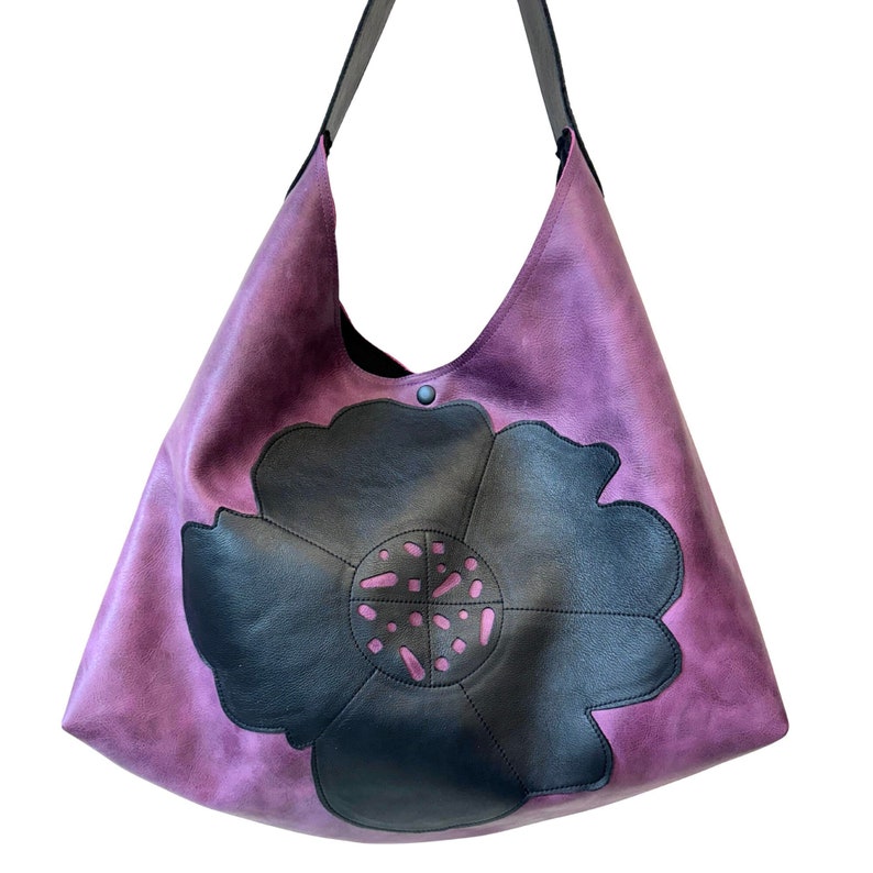 Puede incluir: Un bolso tote de cuero morado con un gran dise&ntilde;o de flor negra. La flor tiene un centro rosa y est&aacute; hecha de cuero.