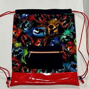 May include: Drawstring backpack with a black top and a red glittery bottom. The main body features a colorful print with various spooky characters. A black zippered pocket is on the front, and red cords serve as the drawstring.