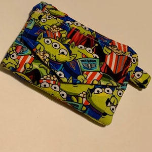 May include: A small, folded fabric wallet featuring a repeating pattern of green aliens with three eyes, holding popcorn and wearing Space Ranger uniforms. The background is blue with red and white striped popcorn containers.