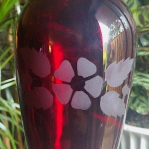 Peut inclure: Un vase en verre rouge foncé avec une base évasée et un bord festonné. Le vase présente des motifs floraux gravés. Le vase est tenu par une main, avec des plantes vertes en arrière-plan.
