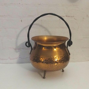 May include: A copper cauldron with a hammered texture and three small legs. It has a black, arched handle. The cauldron is likely used for decorative purposes or for cooking.