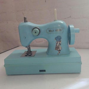 May include: A vintage, pale blue Holly Hobby sewing machine. The machine has a matching base and features a decorative sticker of a girl. The machine has a hand wheel, a needle, and a spool holder.