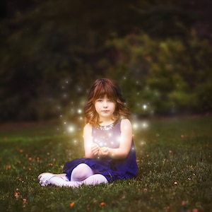 May include: A young girl in a blue dress sits in a grassy field, holding her hands out in front of her.  She is surrounded by small, glowing lights.