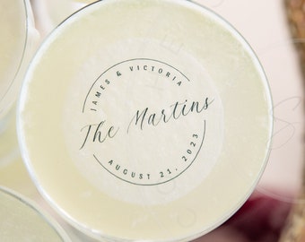 May include: A round, white cocktail topper featuring black text that reads James & Victoria, The Martins, and August 21, 2023, arranged in a circular design. The topper is placed on the surface of a pale yellow liquid in a glass.