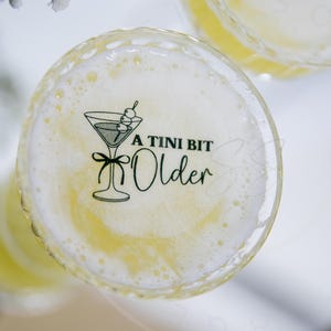 May include: A clear glass with a yellow liquid and a sticker that says "A Tini Bit Older" with a martini glass graphic.
