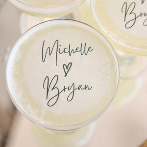 May include: A round cocktail topper made of thin, edible paper featuring the dark green printed text Michelle and Bryan with a small heart symbol in the center. The topper is placed on the surface of a pale yellow liquid in a glass.