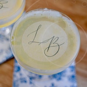 May include: Overhead view of two cocktails in stemmed glasses. The drinks have a pale yellow hue and are topped with a foamy layer. Each glass has a circular sticker with the cursive letters "LB". The glasses rest on blue and white patterned napkins.